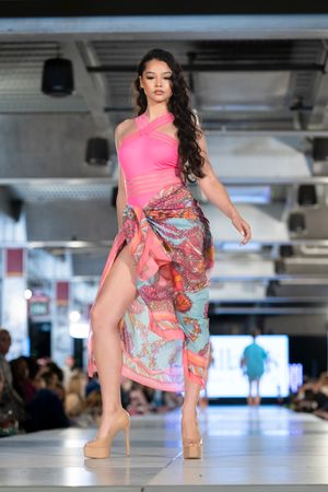 Model walks runway in pink one-shoulder dress with colorful paisley wrap skirt and nude heels.