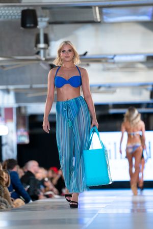 Model walks runway in blue bikini top and striped palazzo pants with turquoise beach bag.