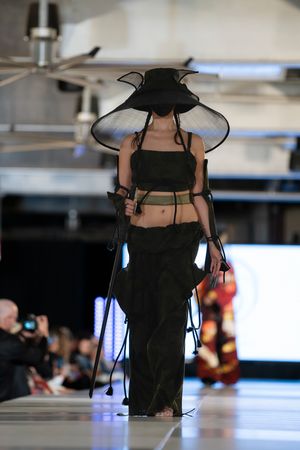 Fashion model walks runway in dramatic black ensemble with wide-brimmed hat during indoor fashion show.