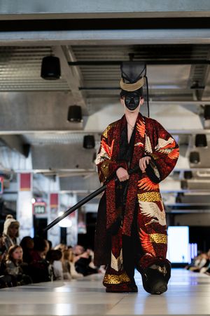 Model showcases vibrant red and gold patterned coat with black accessories on illuminated catwalk.