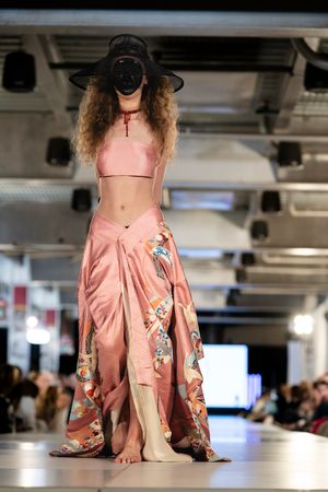 Pink floral dress with asymmetrical hem shown on fashion runway with dramatic lighting and movement.