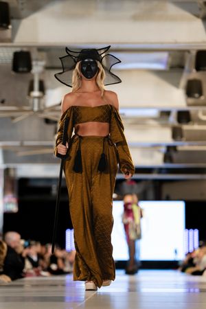 Fashion model wearing brown off-shoulder dress and black hat walks runway in industrial-style venue.