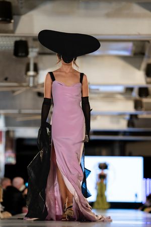 Model wearing pink dress and black gloves poses on illuminated catwalk.