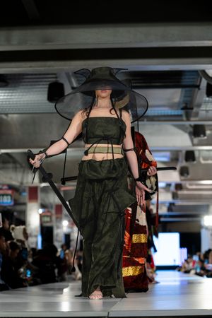 Dark olive dress with dramatic styling presented on illuminated catwalk.