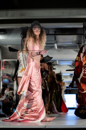 Model walks runway in flowing pink satin dress with dramatic lighting in industrial space.