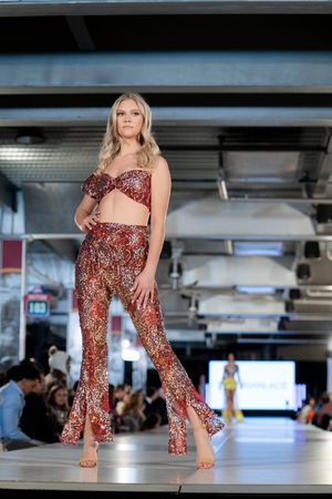 Model walks runway in red patterned two-piece outfit with flared pants.