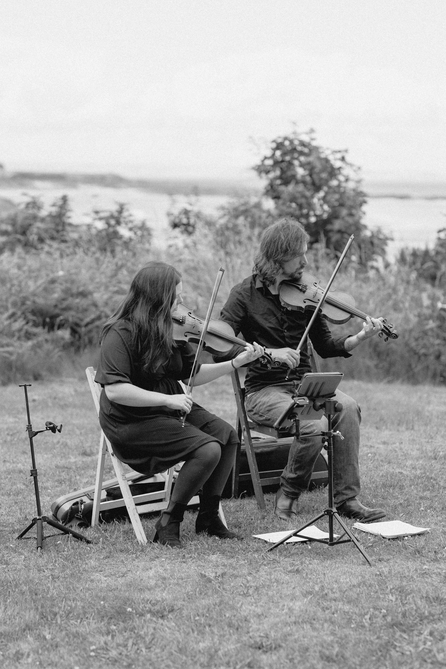 Two musicians perform traditional folk music with violins while seated outdoors on a grassy hillside.