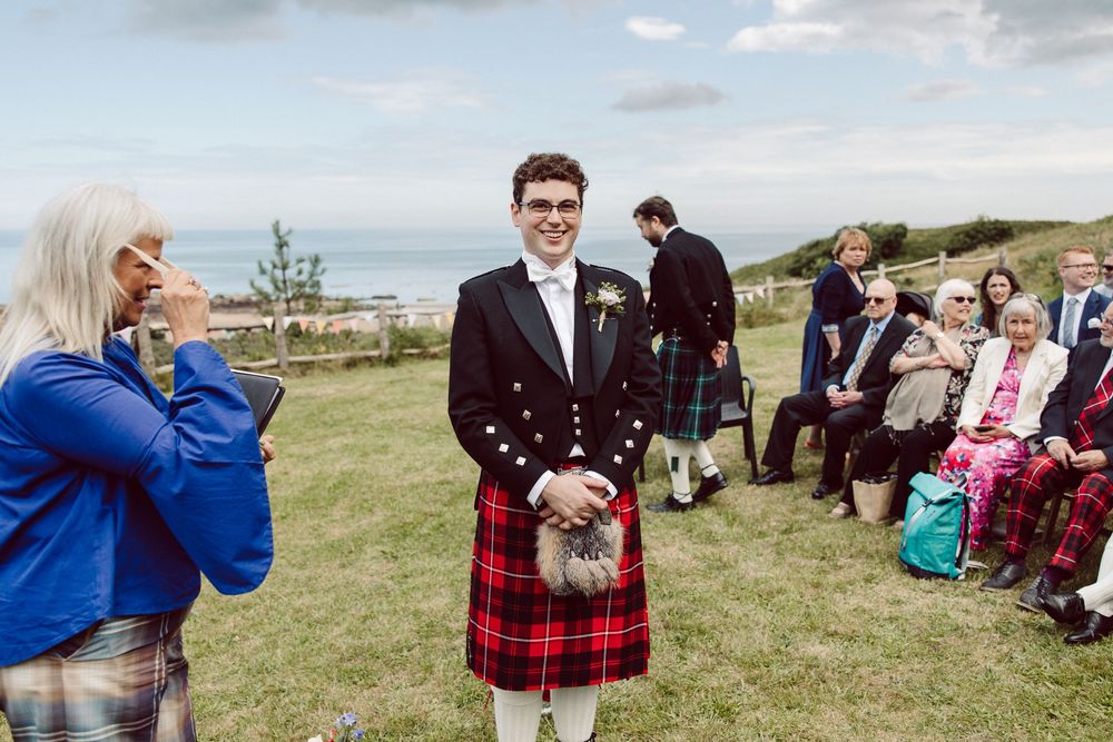 A traditional Scottish wedding celebration outdoors with guests and someone wearing a red tartan kilt on a grassy hillside.
