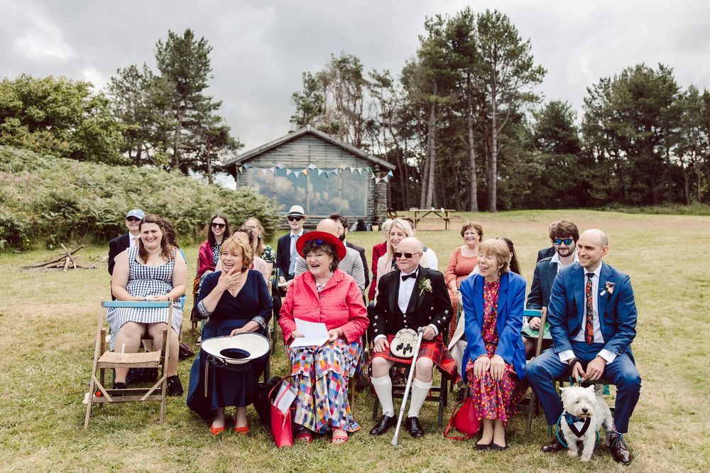 Wedding party members sit together on rustic wooden benches in a scenic outdoor garden setting.