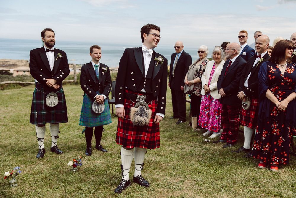 Wedding guests in traditional Scottish kilts and formal attire gather outdoors on grassy hillside for ceremony.