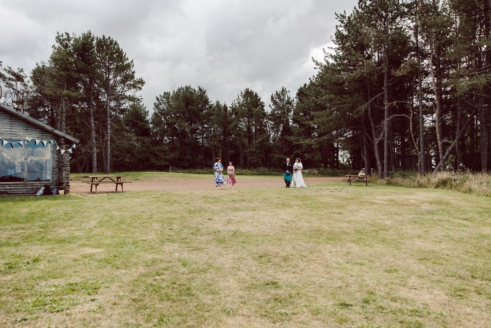 People gather on a grassy field surrounded by tall pine trees on an overcast day.