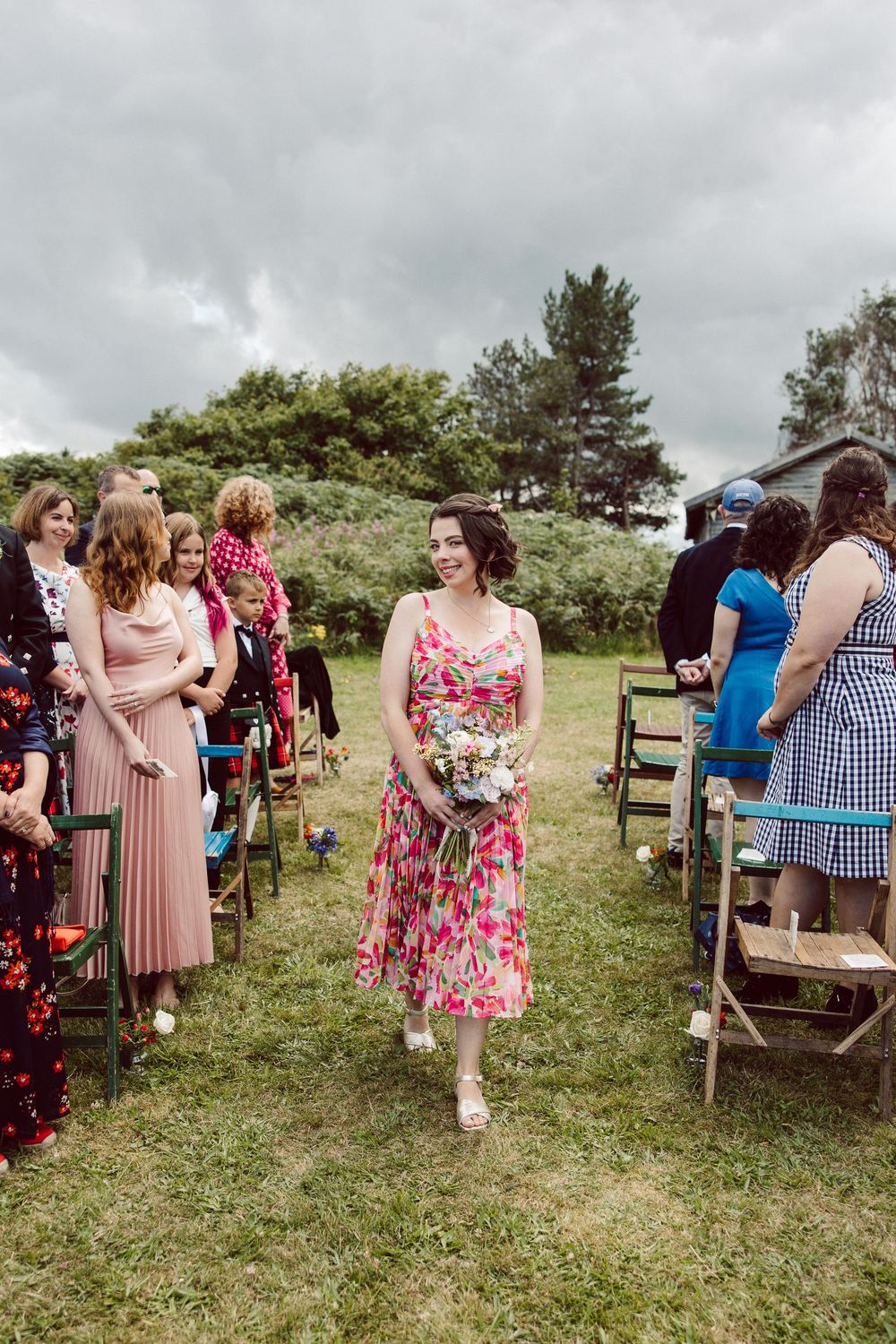 Wedding guests in colorful floral dresses gather at an outdoor ceremony with wooden chairs on grassy terrain.