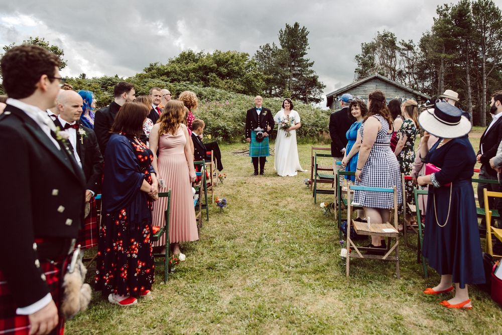 Outdoor Scottish wedding ceremony setup with guests seated on wooden chairs on a grassy hillside.
