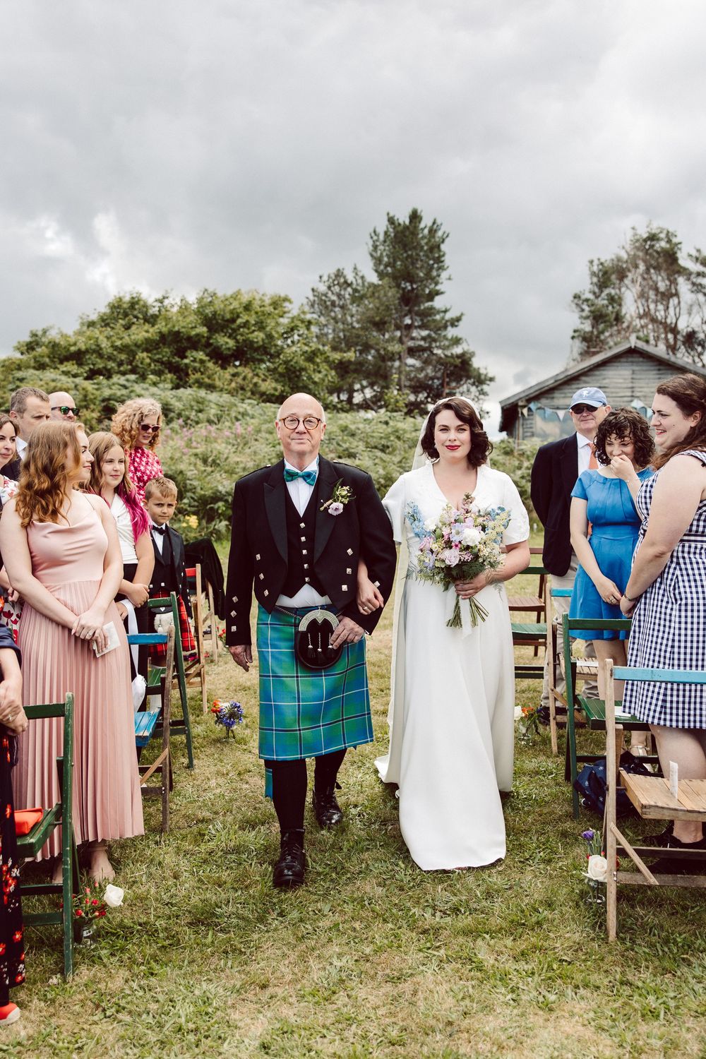 Wedding party and guests mingling on a green hillside during outdoor Scottish-themed celebration.