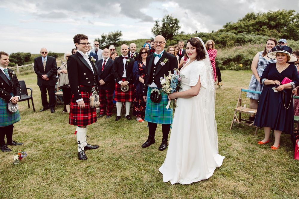 Wedding guests gather outdoors on a grassy hillside for a scenic ceremony with traditional Scottish kilts and formal attire.