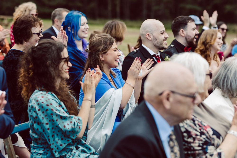 Wedding guests react emotionally during an outdoor ceremony while seated in white chairs.