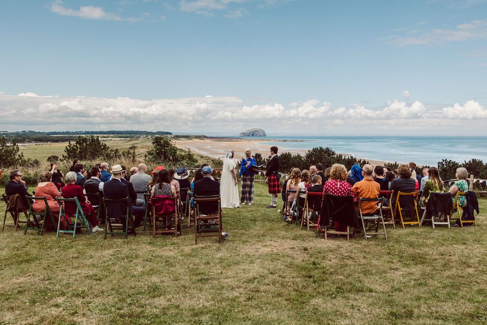 Panoramic view of outdoor wedding ceremony setup with chairs on grassy field overlooking coastal scenery.