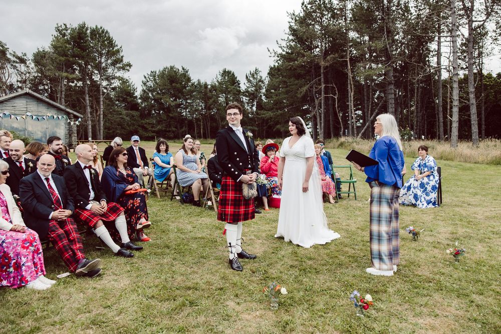 Panoramic view of an outdoor wedding ceremony with guests seated on wooden chairs surrounded by pine trees.