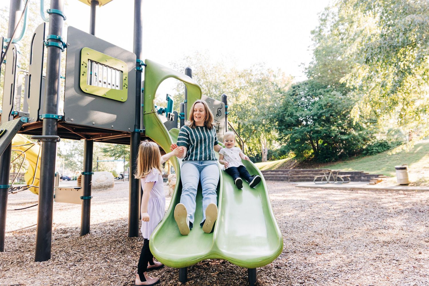 A long, winding playground slide structure stretches across a natural outdoor setting with trees in the background and a mom sliding down
