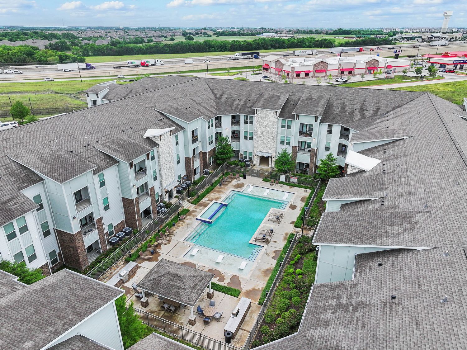 Aerial view of modern apartment complex with outdoor pool, patio seating, and highway views.