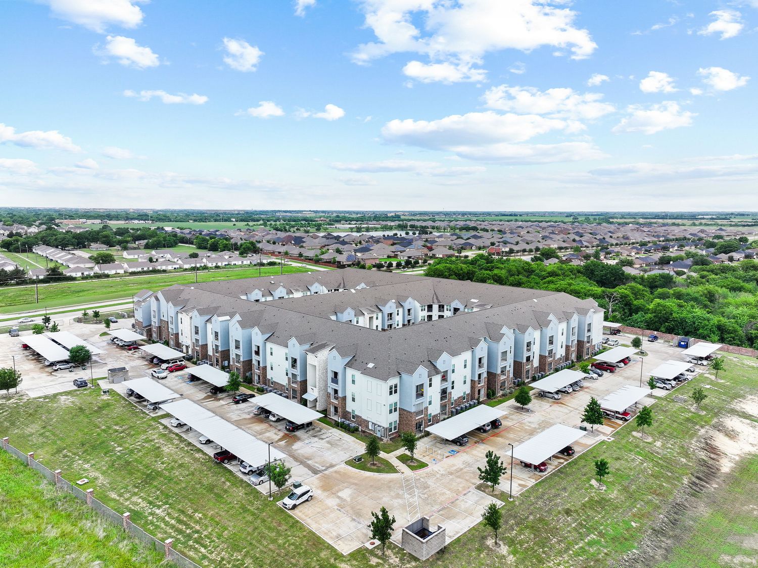 Aerial view of a modern apartment complex with parking lots surrounded by suburban neighborhood under a partly cloudy sky.