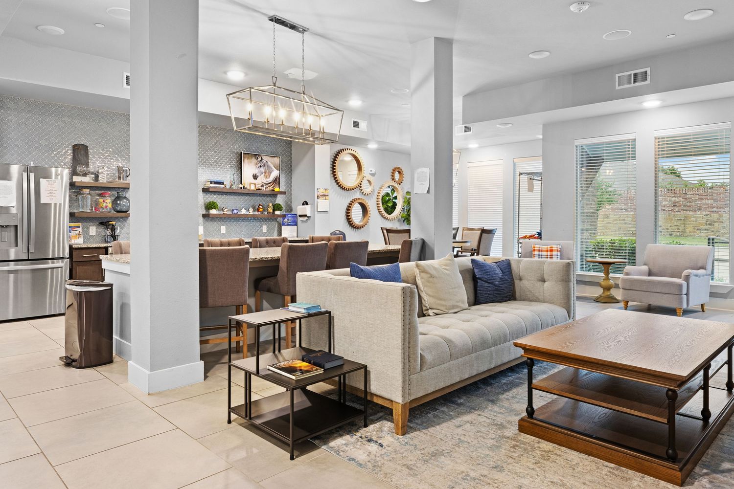 Modern open-concept living room with gray sofa, chandelier, tile floors, and open kitchen in a bright, elegant space.