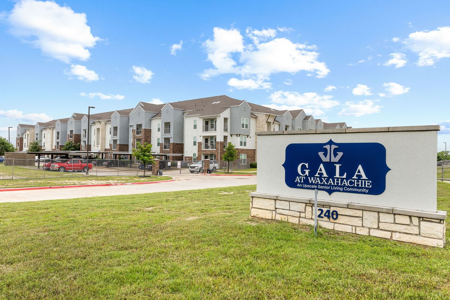 Sign for Gala at Waxahachie apartment community with multi-story residential buildings and green lawn in background.