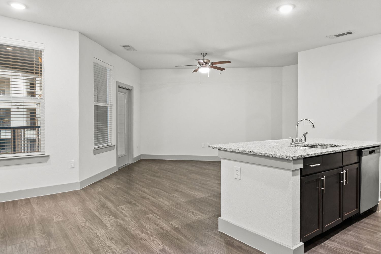 Modern kitchen island with granite countertop and sink, dark cabinets, hardwood floors, and open living area with ceiling fan.