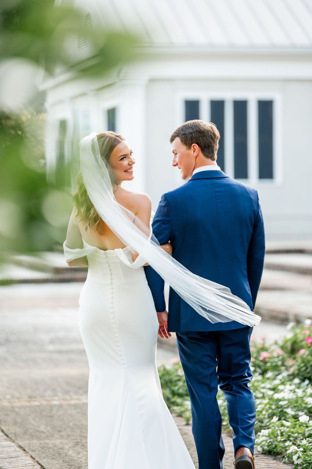 Newlywed couple walking together with bride in white dress and groom in navy suit along garden path.