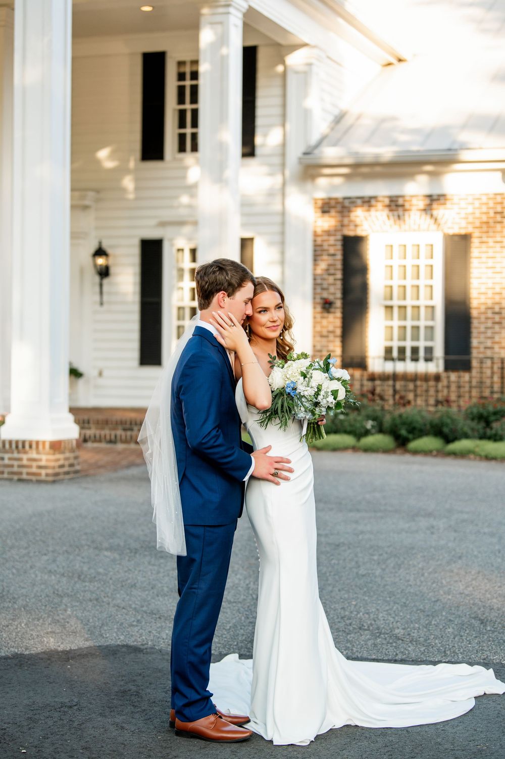 Elegant wedding couple embraces on grand white columned porch with trailing bridal veil.