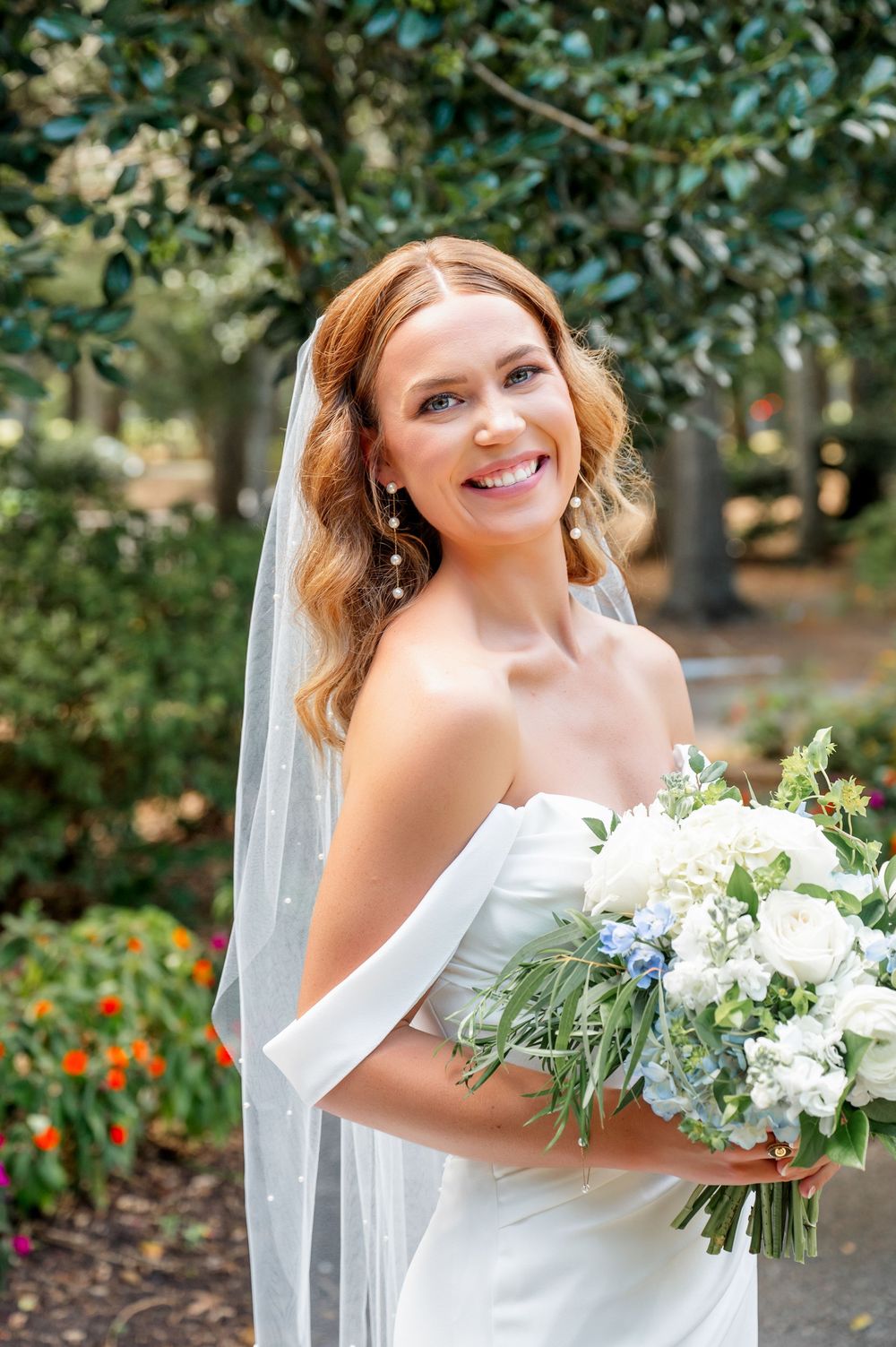 Bride in white off-shoulder gown holds bouquet of white flowers against garden backdrop.