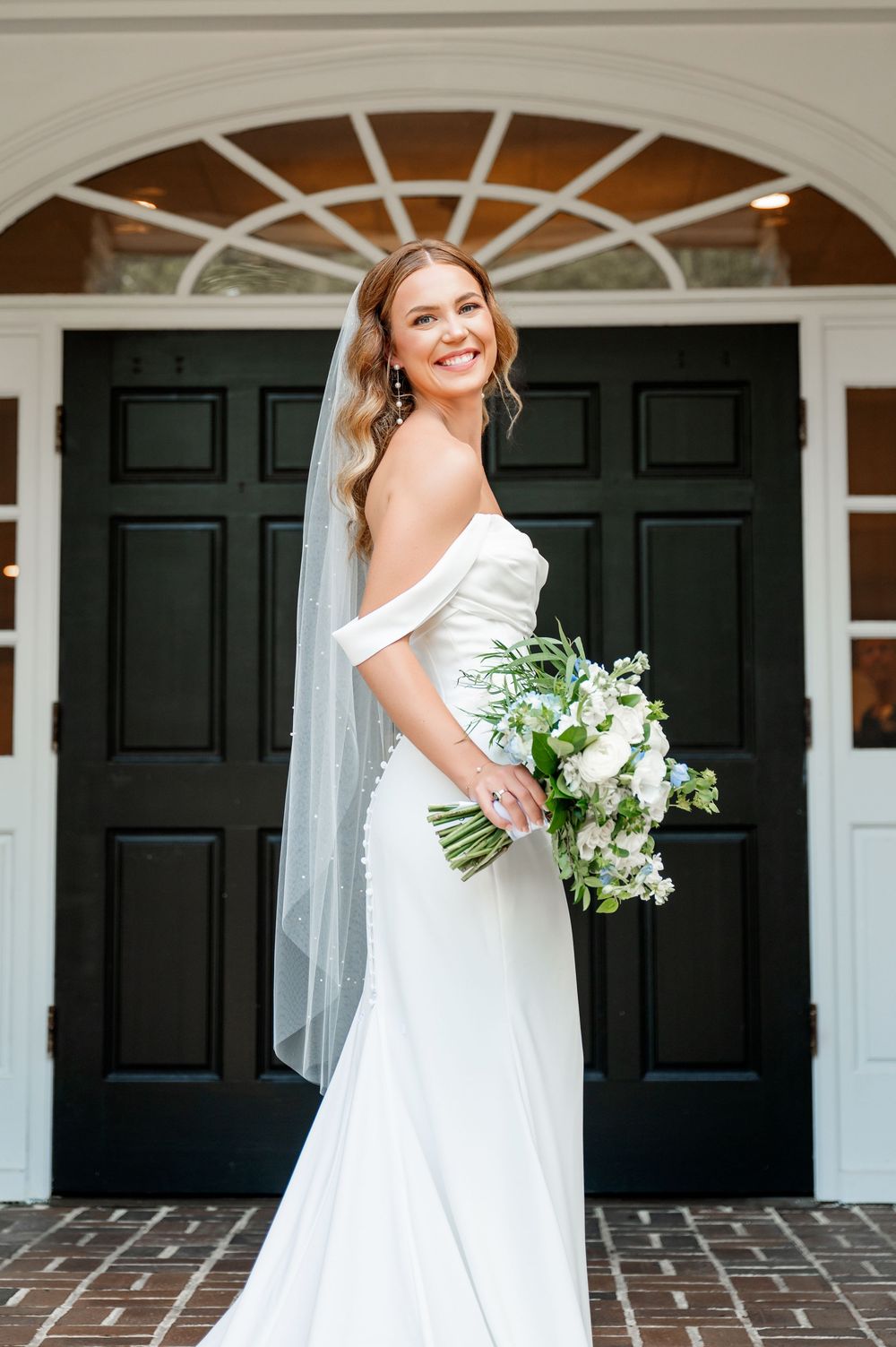 Bride poses in elegant white gown with veil against black doors and white columns.