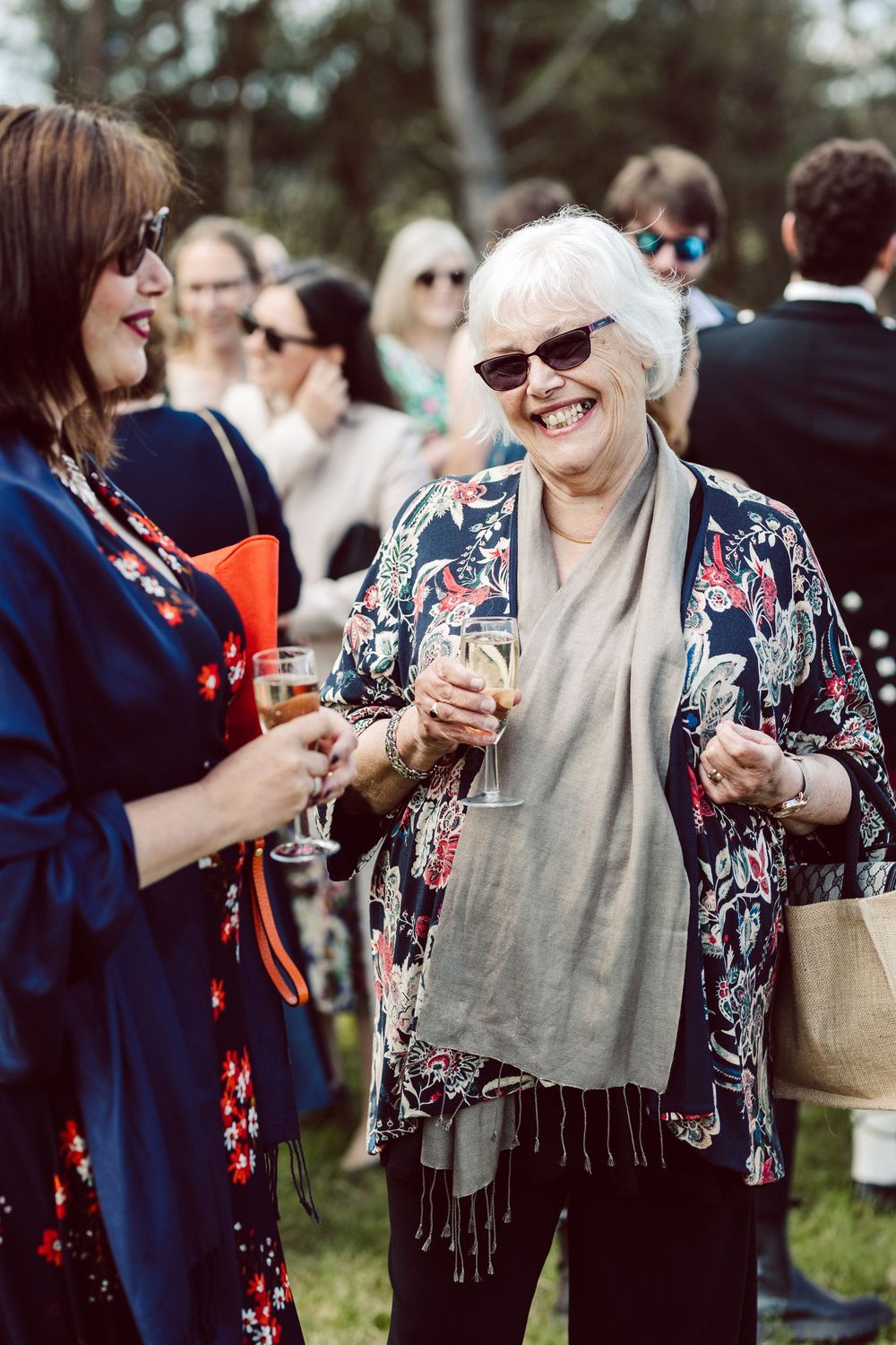 Two women enjoying drinks and conversation at an outdoor social gathering, one wearing a floral kimono and sunglasses.