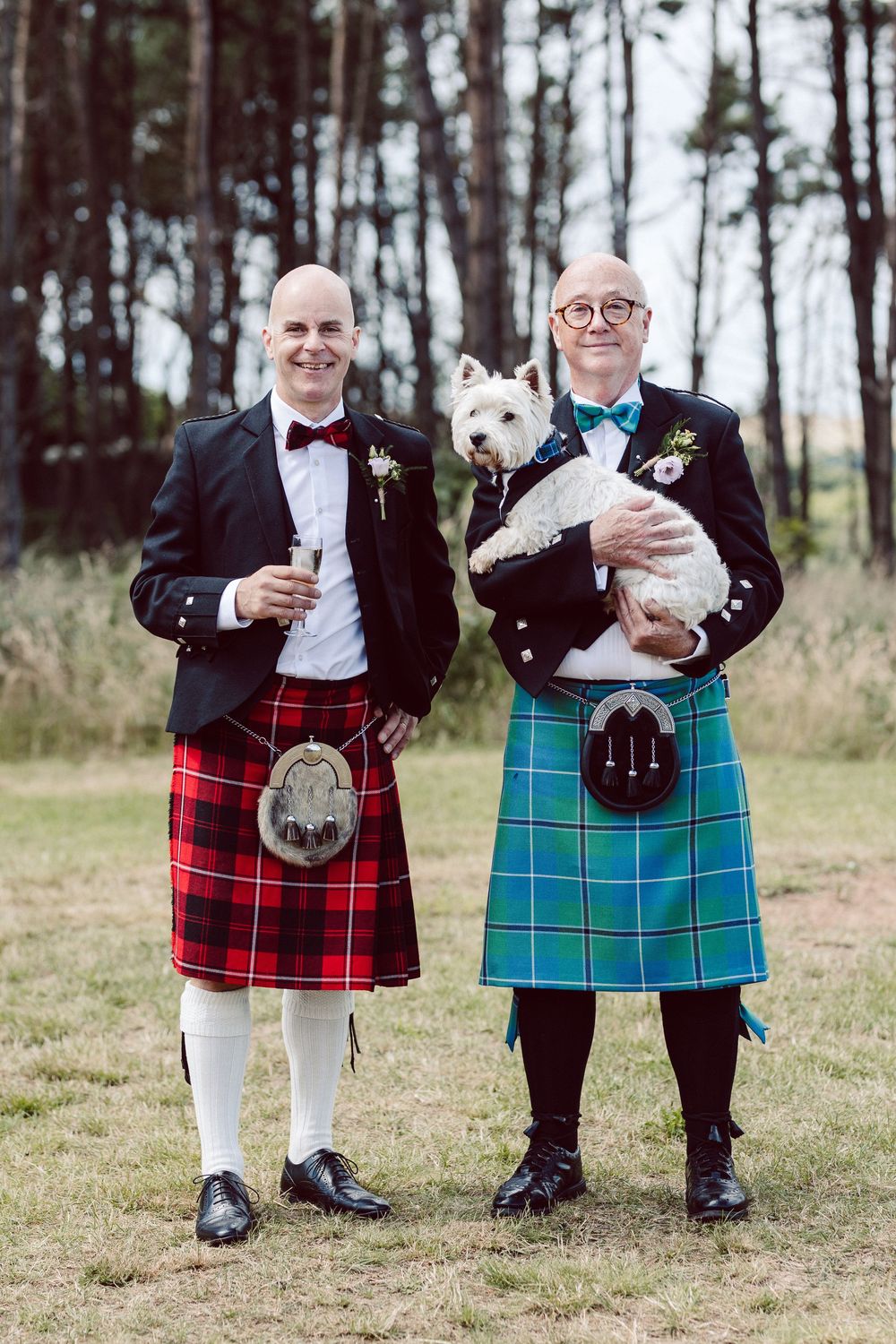 Two people in kilts holding small white dogs while standing in a grassy field with trees in the background.