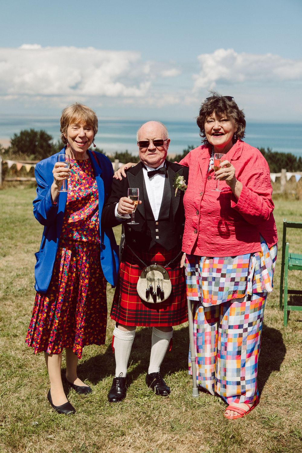 Three people stand together in a sunny field wearing colorful dresses and formal attire while holding drinks.