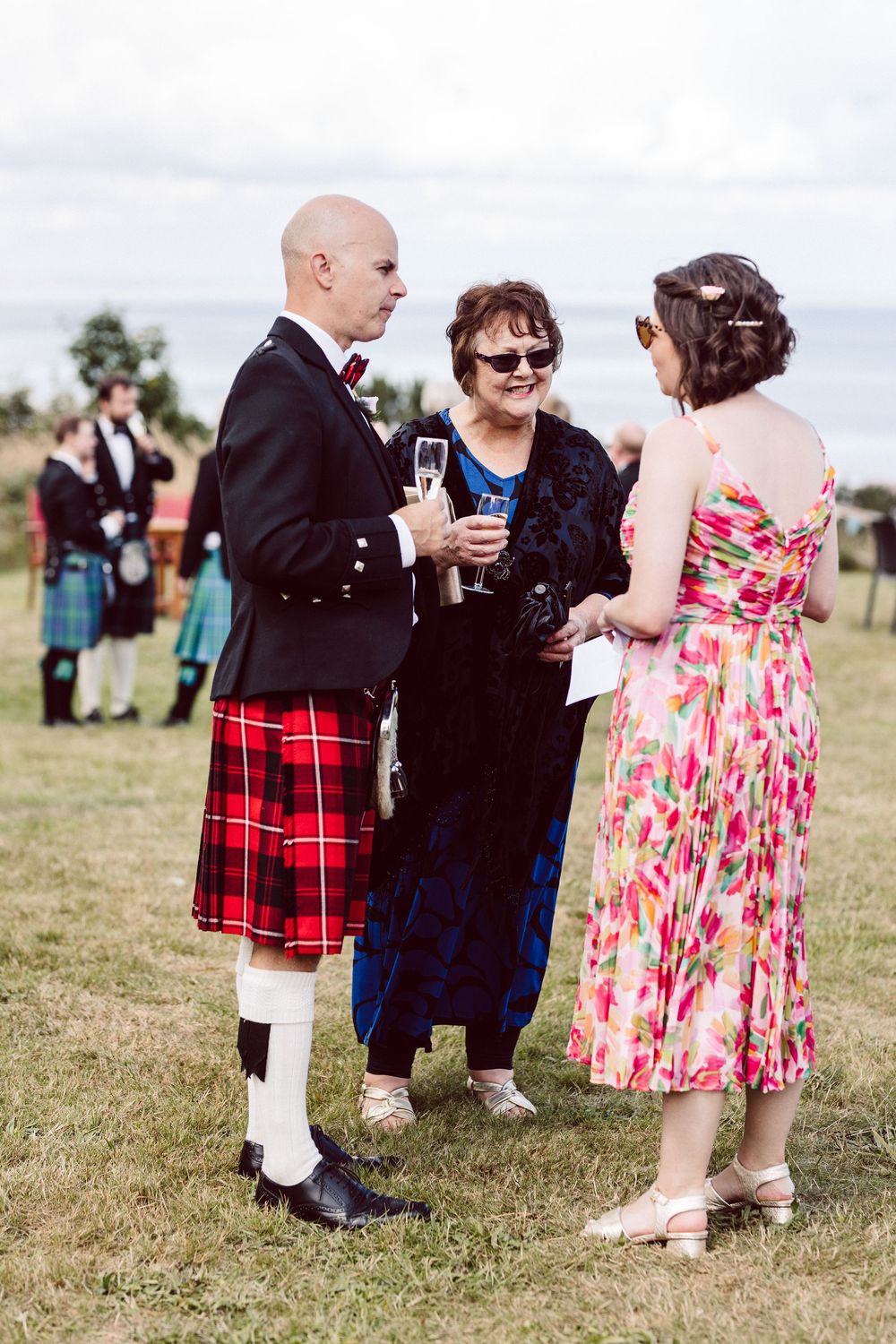 Wedding guests in Scottish kilts and floral dresses mingle on grassy lawn during outdoor reception.