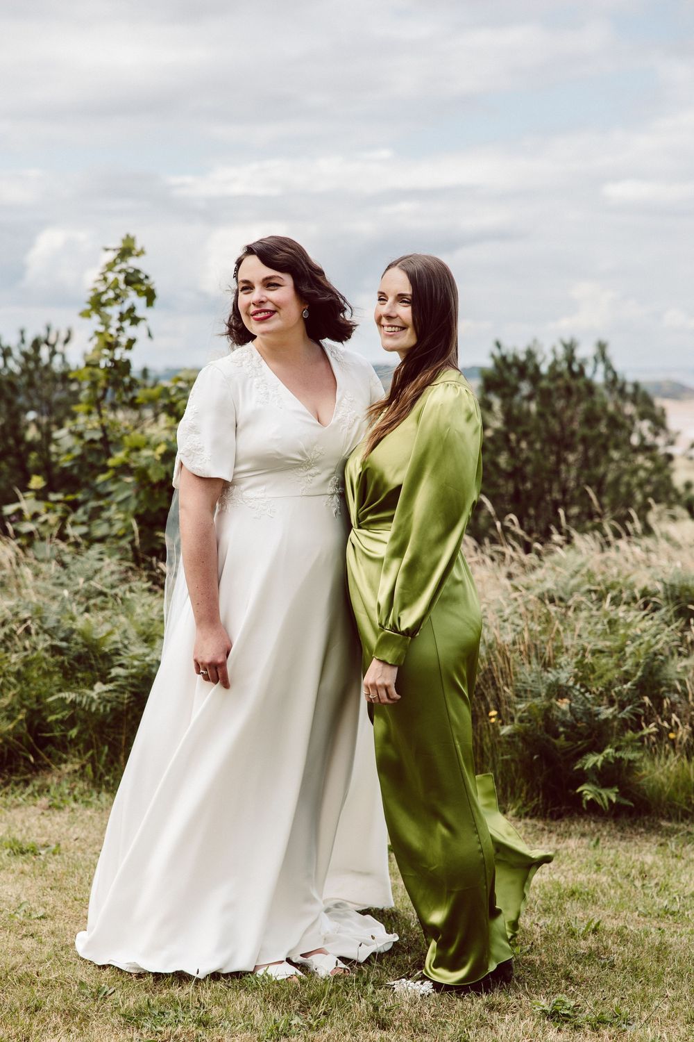 Two friends in white and green dresses share a candid moment laughing together on a grassy hillside.