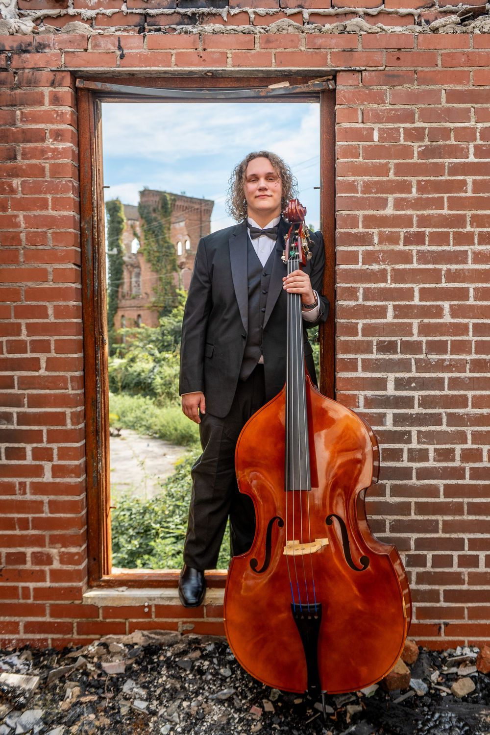 Professional musician poses with upright bass against rustic brick wall in formal attire.