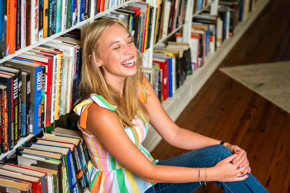 A person laughs while sitting in a library surrounded by colorful bookshelves.