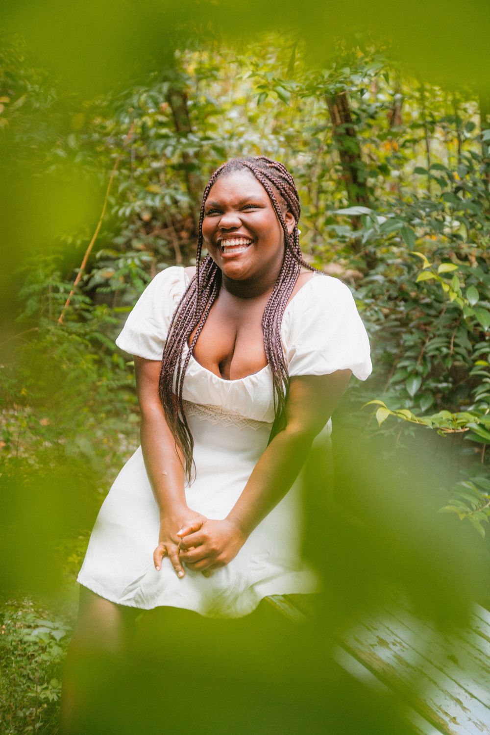 A joyful person in a white dress poses outdoors among lush green foliage in a natural setting.