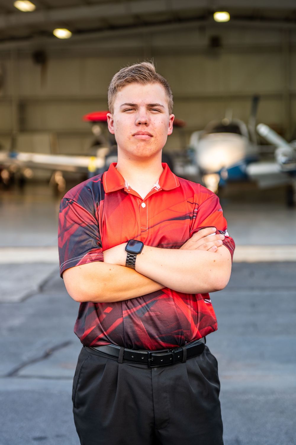 Person in red and black uniform stands with arms crossed in parking garage.