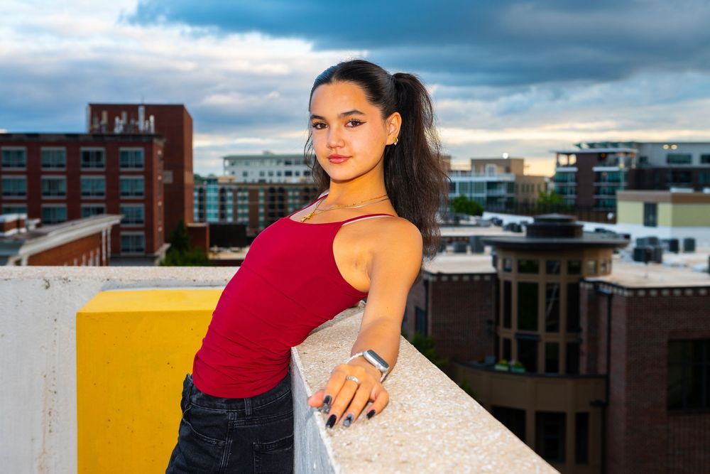 Person in casual attire poses on urban rooftop with city buildings and dramatic cloudy sky in background.