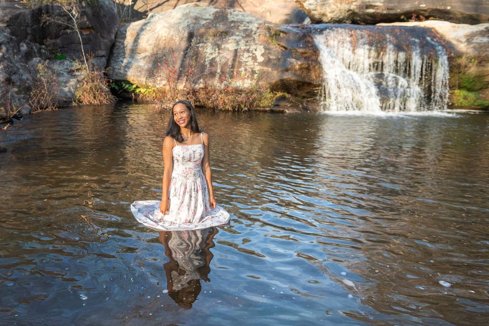 A person in a white dress poses in a scenic waterfall pool surrounded by rocky cliffs and autumn foliage.