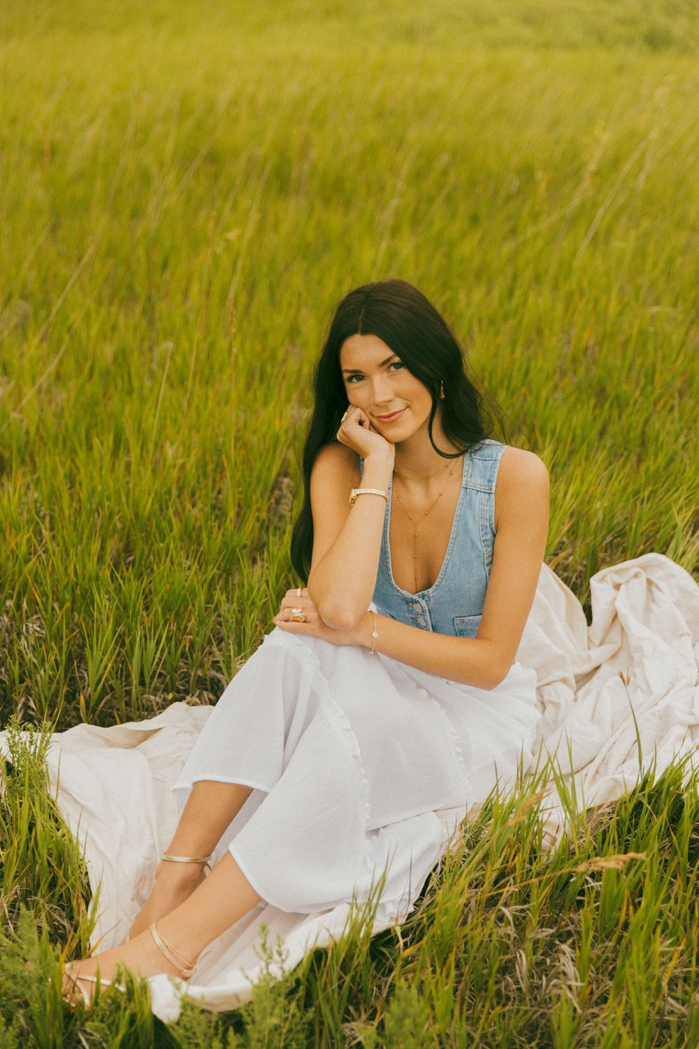 Woman in white dress and denim vest lounging in tall grass field.