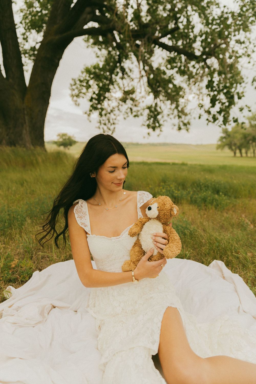 Sweet moment with a teddy bear under sprawling oak trees in a white flowing dress.