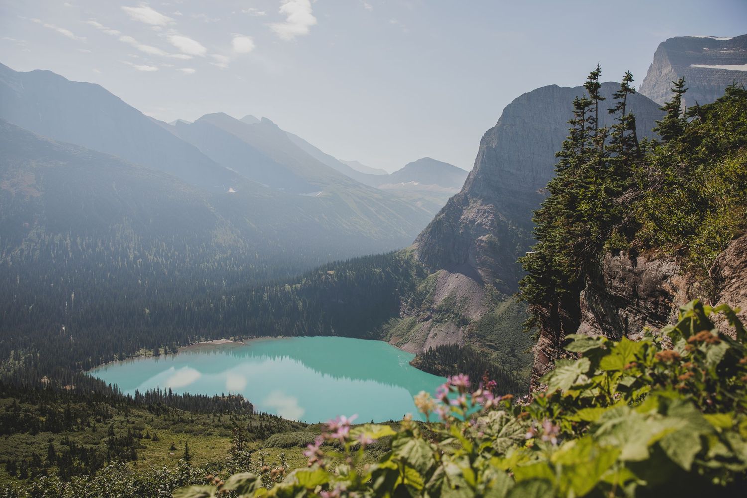 Panoramic mountain landscape with turquoise alpine lake surrounded by rugged peaks and evergreen forest in misty conditions.