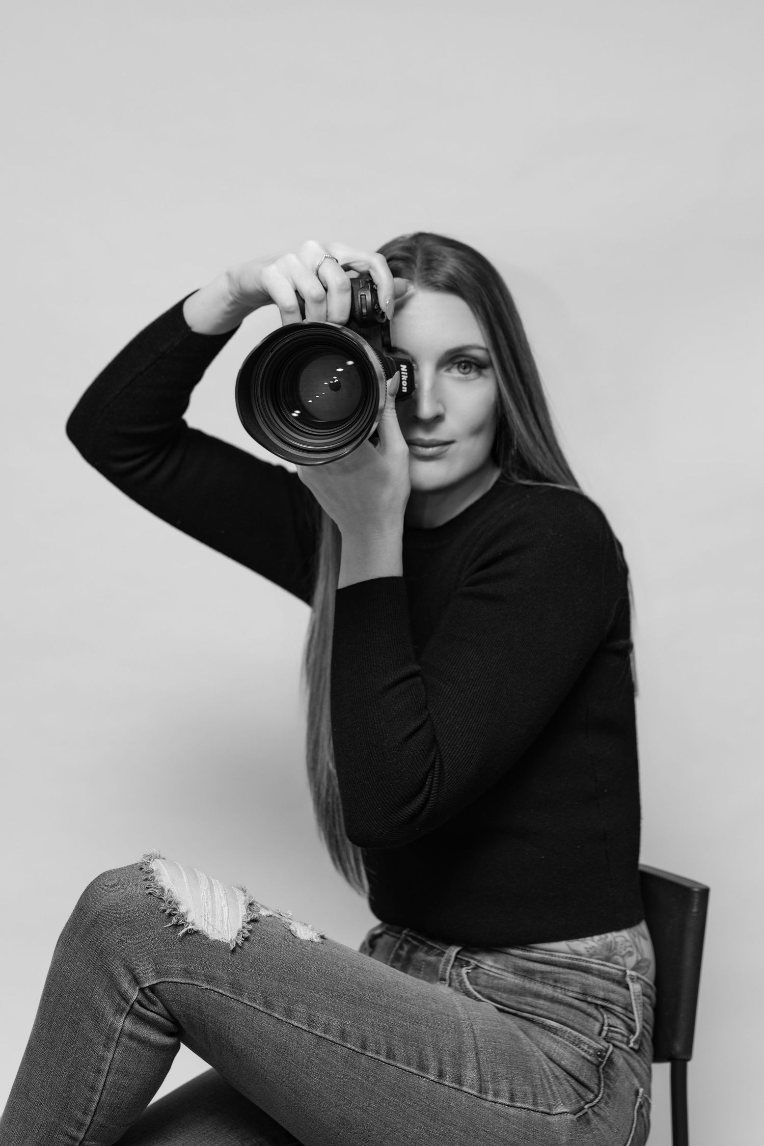 Professional photographer in black turtleneck and jeans poses with camera against beige background.
