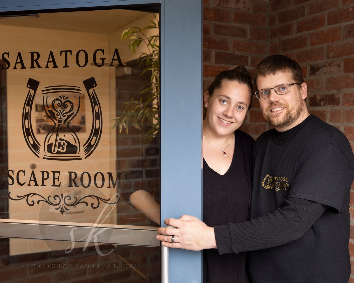 Owners of Saratoga Escape Rooms, Tess and Brian Goodge, smiling at the door of their business in Saratoga Springs.