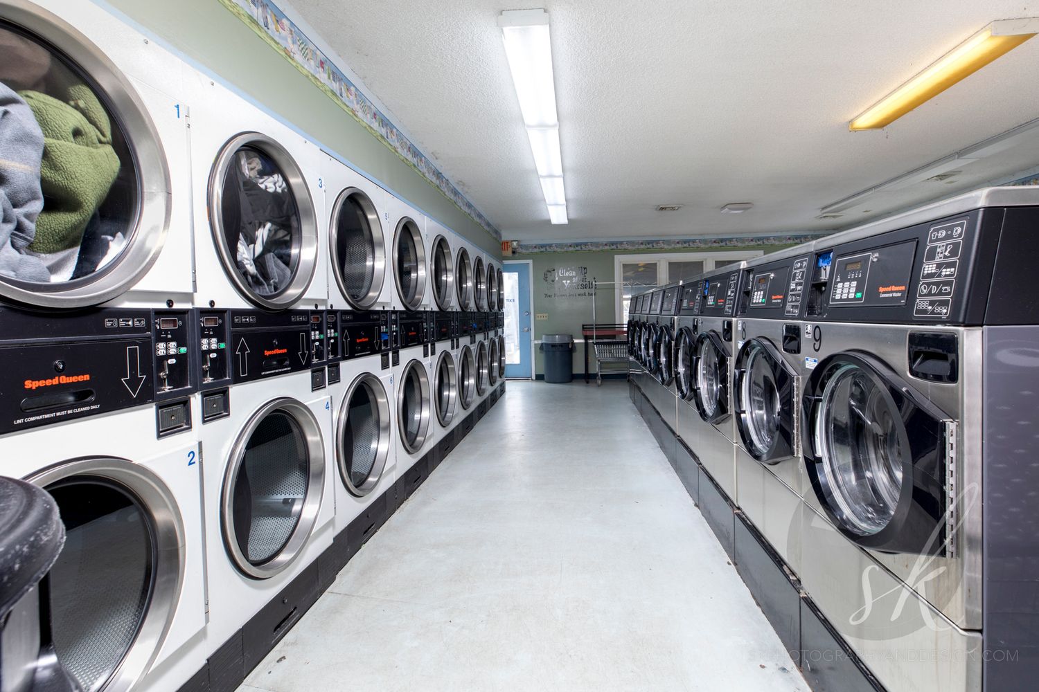 Commercial photo of rows of washers and dryers lining both walls of a clean, bright laundromat in Queensbury.