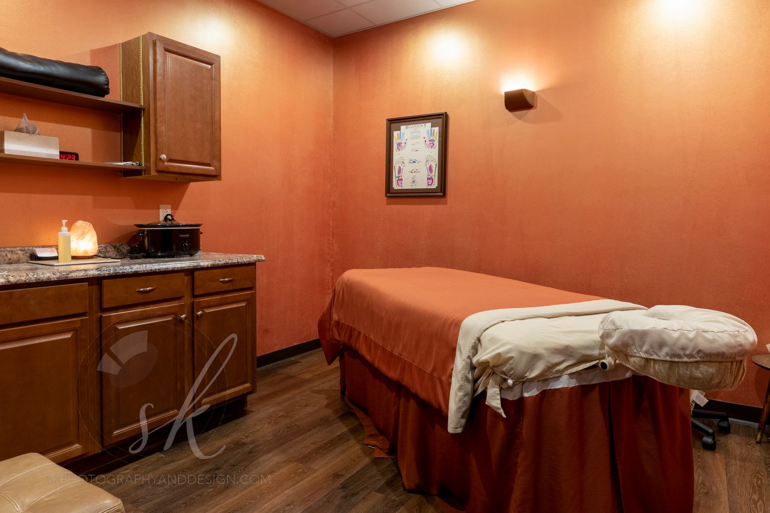 Serene massage therapy room with orange walls, a treatment table with white linens, and wooden cabinetry. Queensbury Spa.